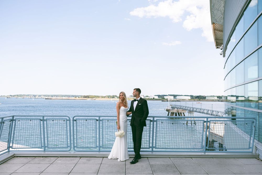Bride and groom pose on a waterfront terrace with scenic harbor views on their wedding day.