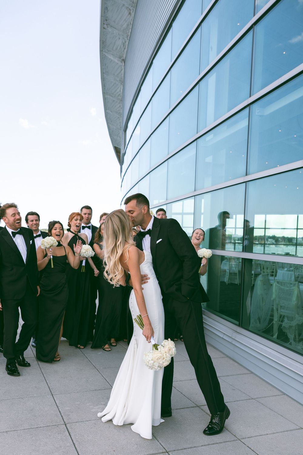 Bride and groom share a kiss while wedding party in black tuxedos and gowns cheers behind them outdoors.