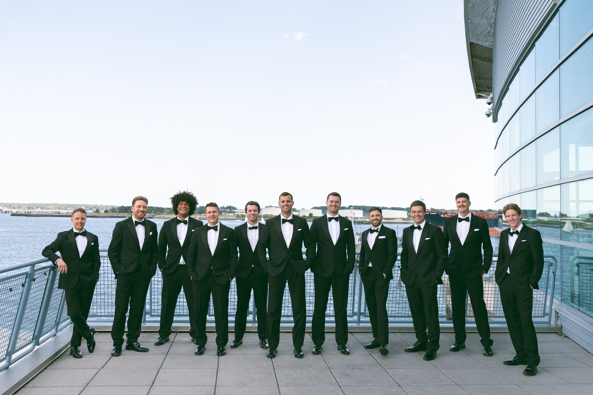 Groomsmen in black tuxedos with bow ties pose on a waterfront venue balcony under a clear blue sky.