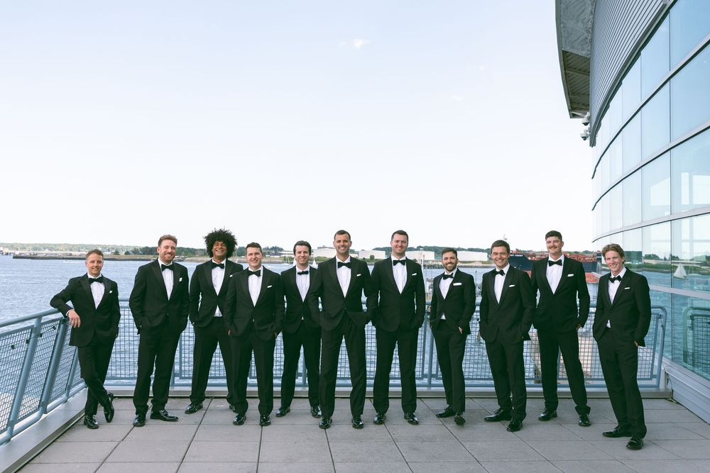 Groomsmen in black tuxedos with bow ties pose on a waterfront venue balcony under a clear blue sky.