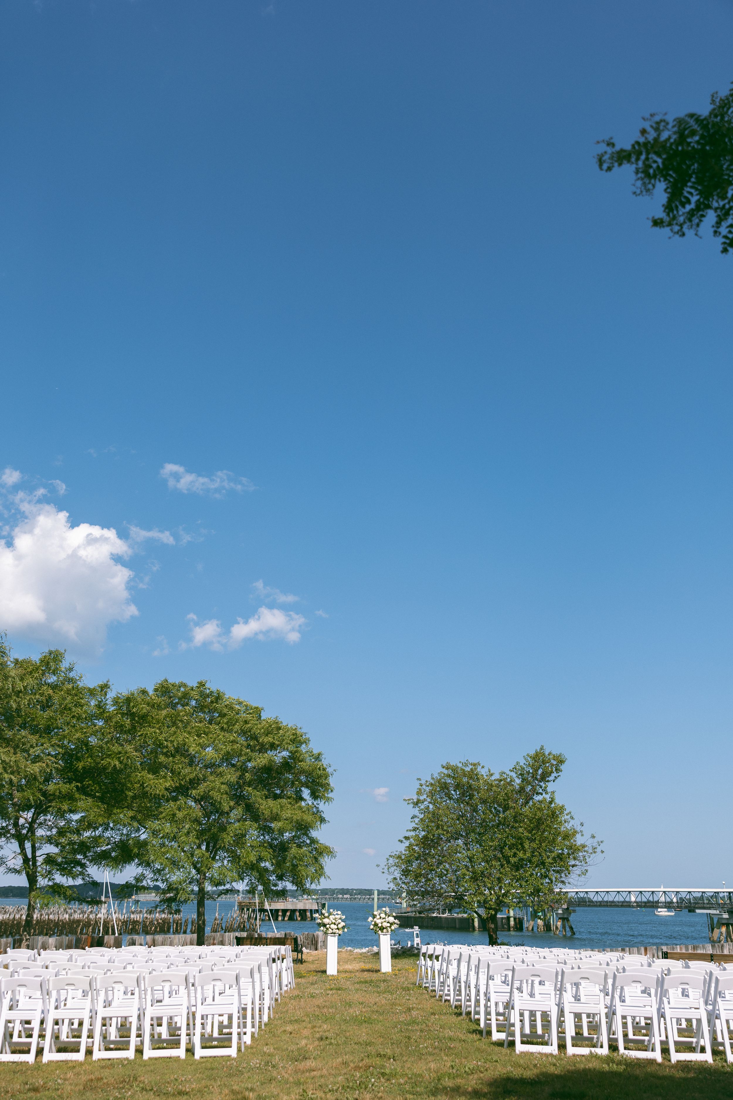Outdoor waterfront wedding ceremony setup with white chairs on green lawn, trees, and blue sky backdrop.