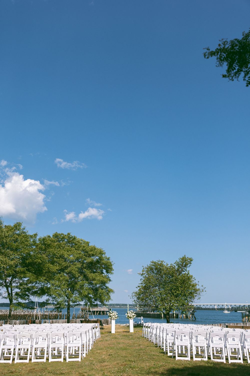 Outdoor waterfront wedding ceremony setup with white chairs on green lawn, trees, and blue sky backdrop.