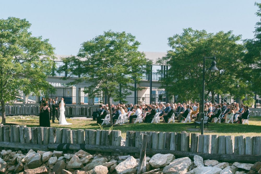 Outdoor wedding ceremony with guests seated on lawn near waterfront, surrounded by trees and modern building backdrop.