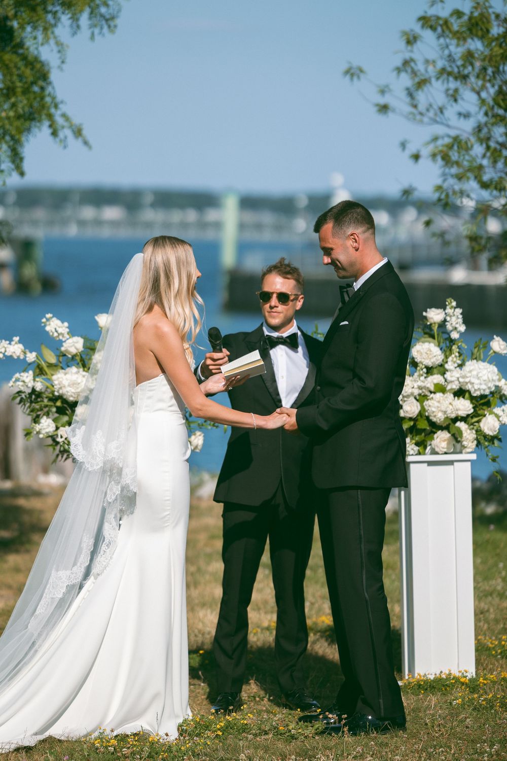 Bride and groom exchange vows outdoors by waterfront with officiant and white floral arrangements on sunny day.