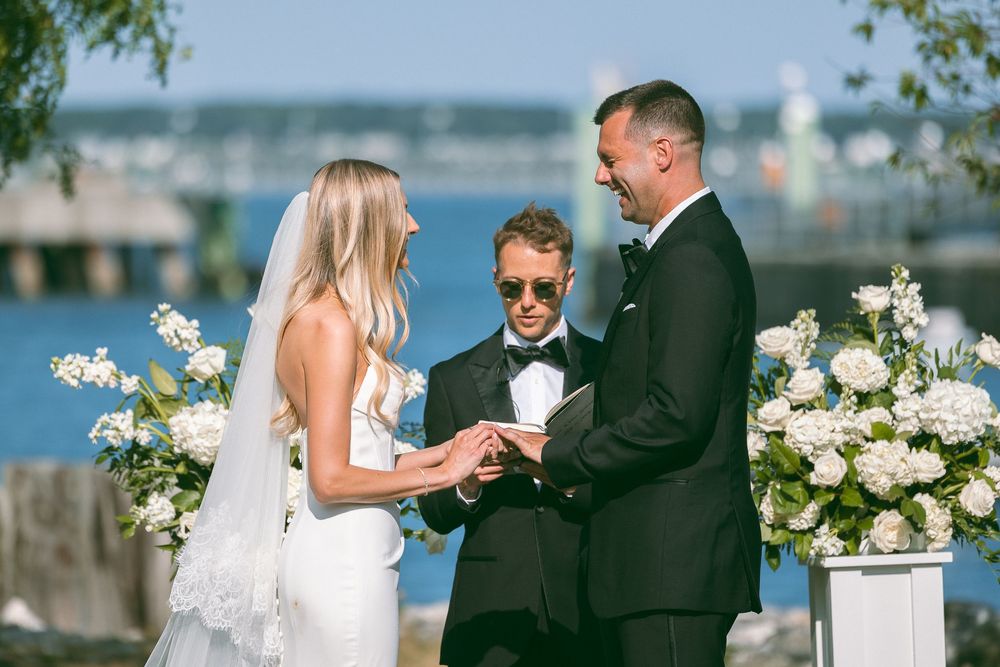 Outdoor wedding ceremony with bride and groom exchanging rings, officiant in sunglasses, white floral arrangements, waterfront backdrop.