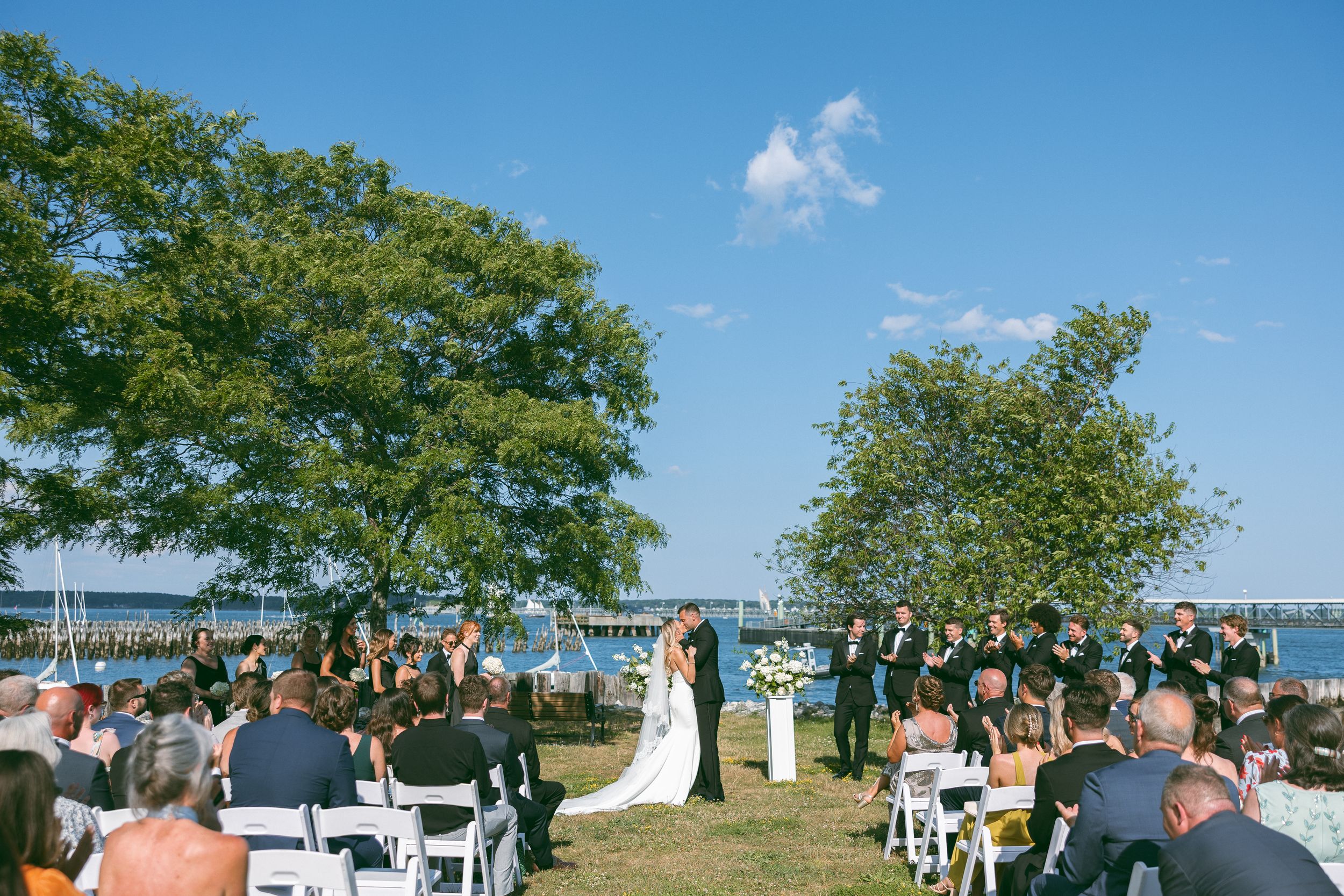 Outdoor waterfront wedding ceremony with guests seated on white chairs under trees on a sunny day.