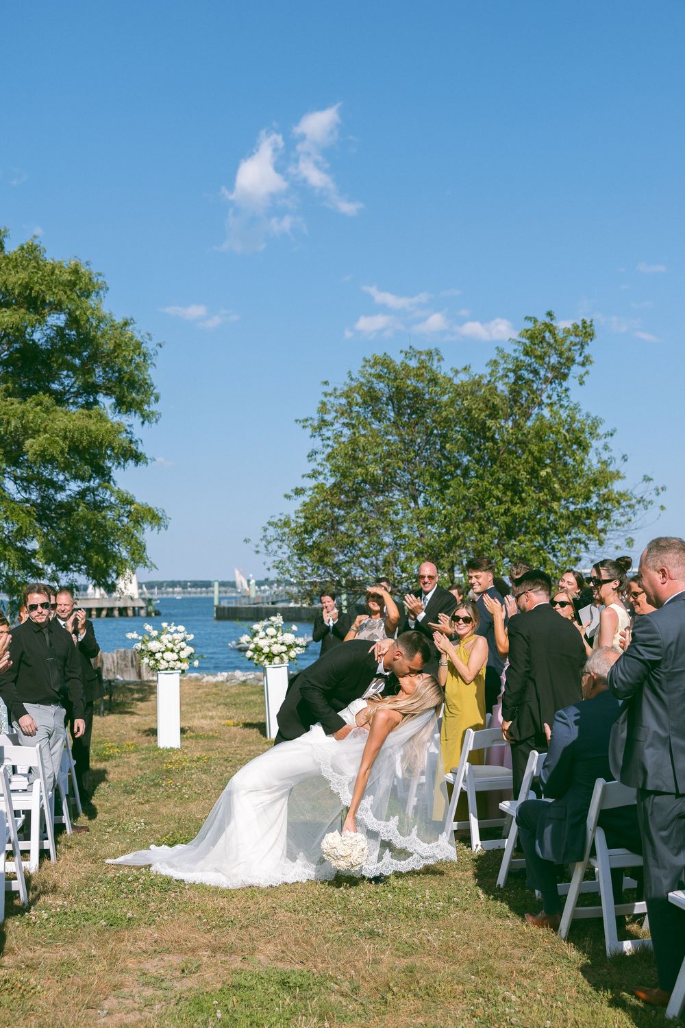 Groom dips bride for a kiss during outdoor waterfront wedding ceremony with guests applauding on a sunny day.