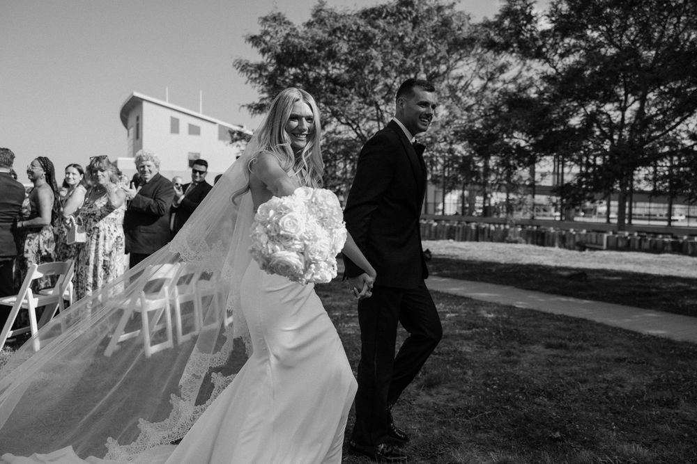 Black and white photo of a smiling bride in a flowing veil walking with a groom in a suit at an outdoor wedding ceremony.