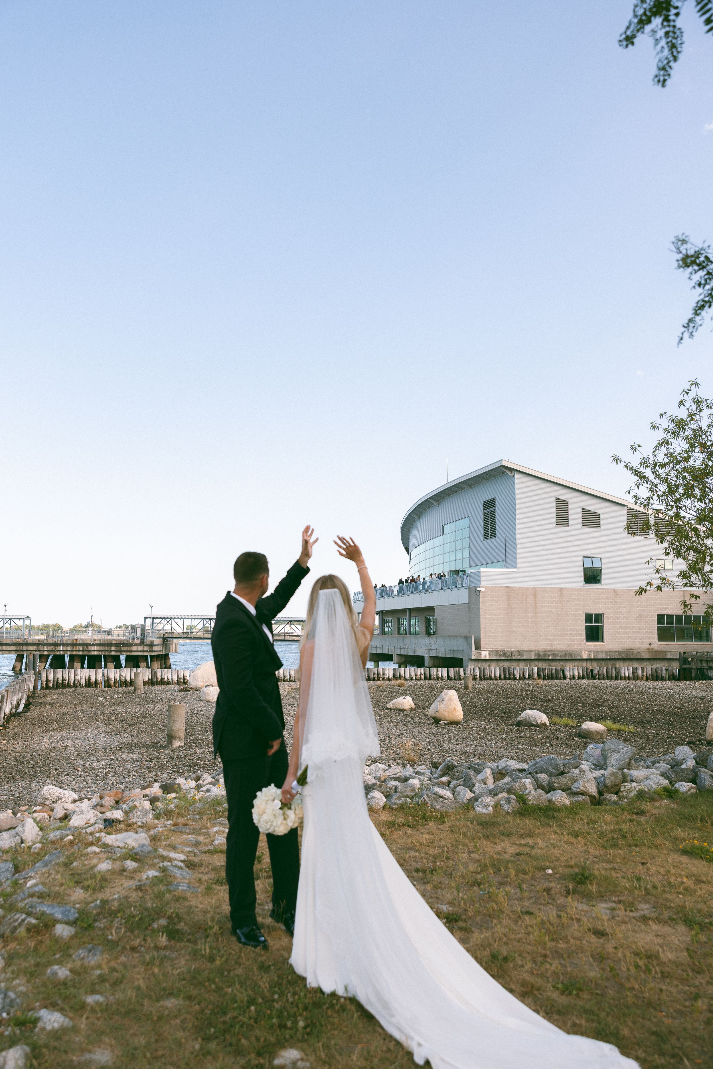 Bride and groom raising hands near waterfront venue, bride in white gown with long veil holding bouquet.
