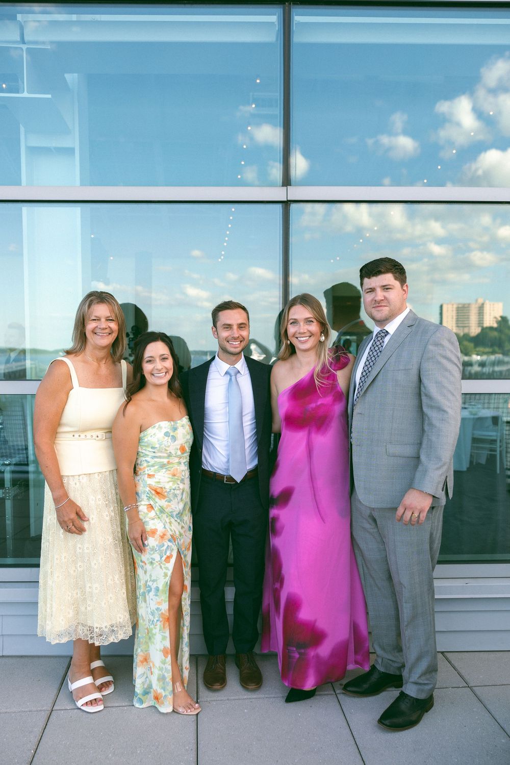 Group of five elegantly dressed people posing together at a formal event in front of large glass windows.