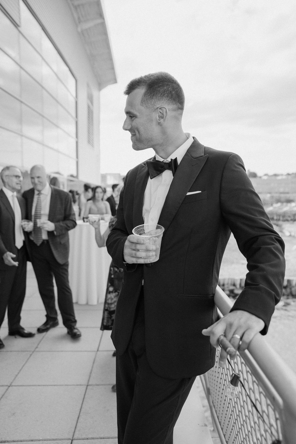 Black and white photo of a groom in a tuxedo holding a drink at an outdoor wedding reception.