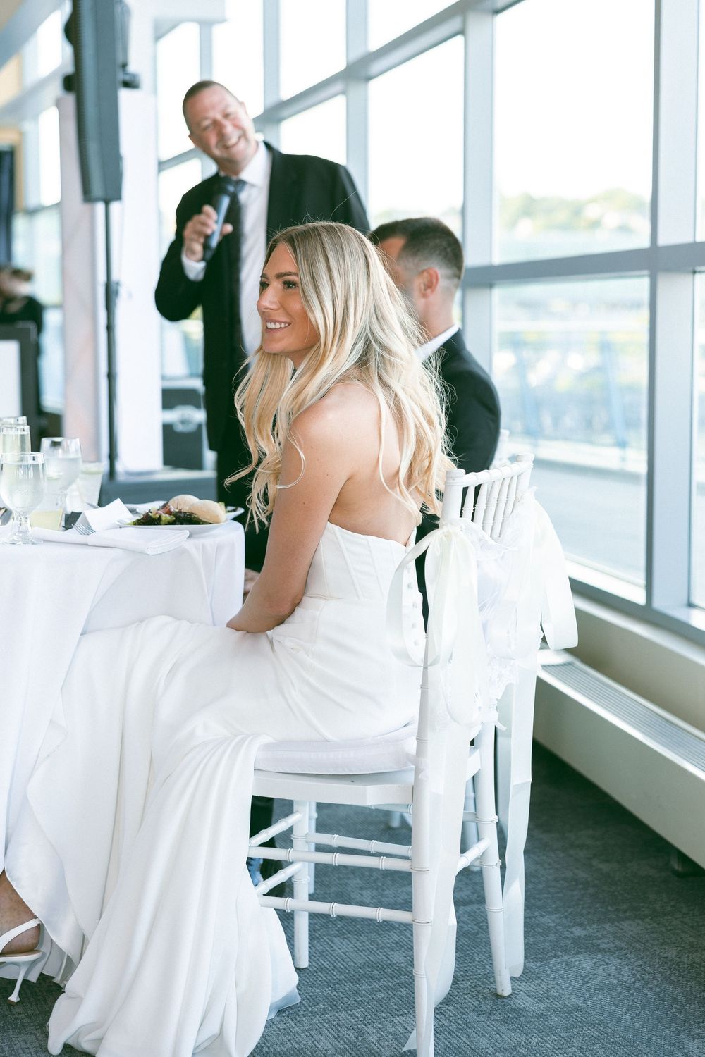 Bride in white gown smiling at wedding reception table while man in suit speaks with microphone behind her.