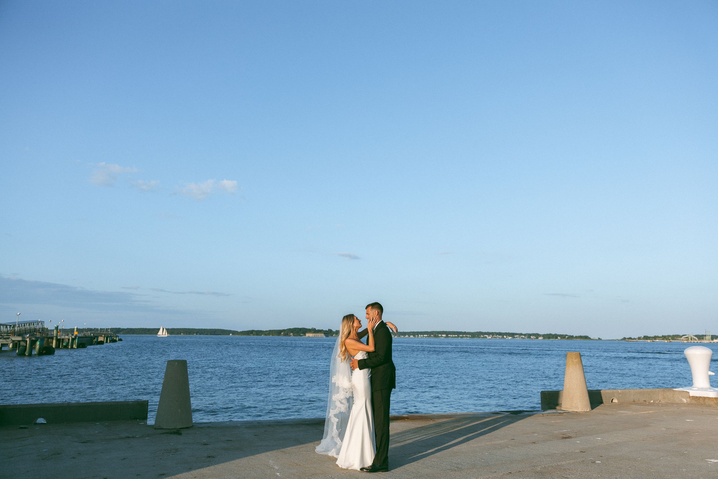 Bride and groom share a romantic embrace on a waterfront dock at sunset with sailboats and blue sky behind them.
