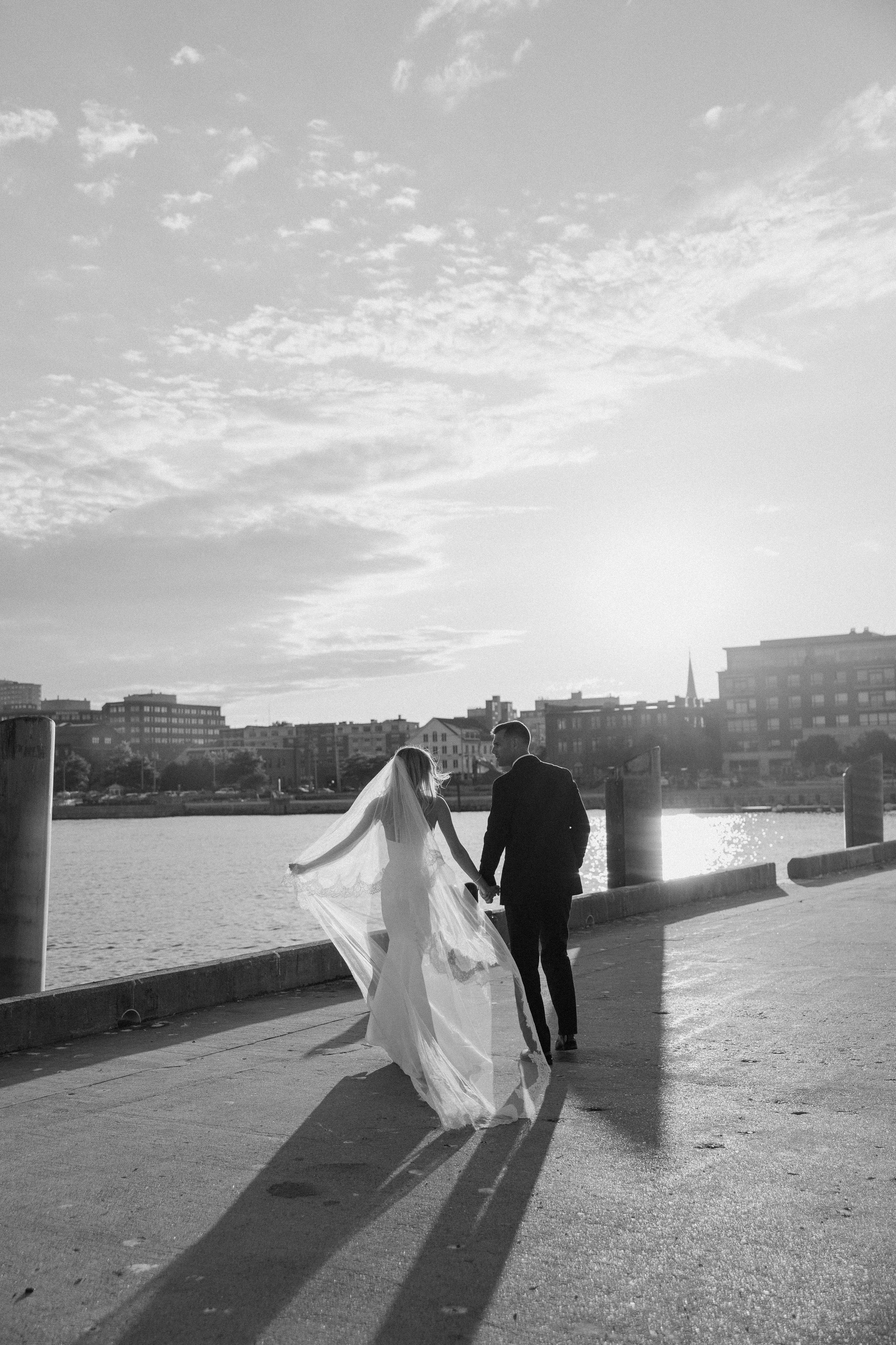 Black and white photo of a bride and groom holding hands, walking along a waterfront promenade at sunset.