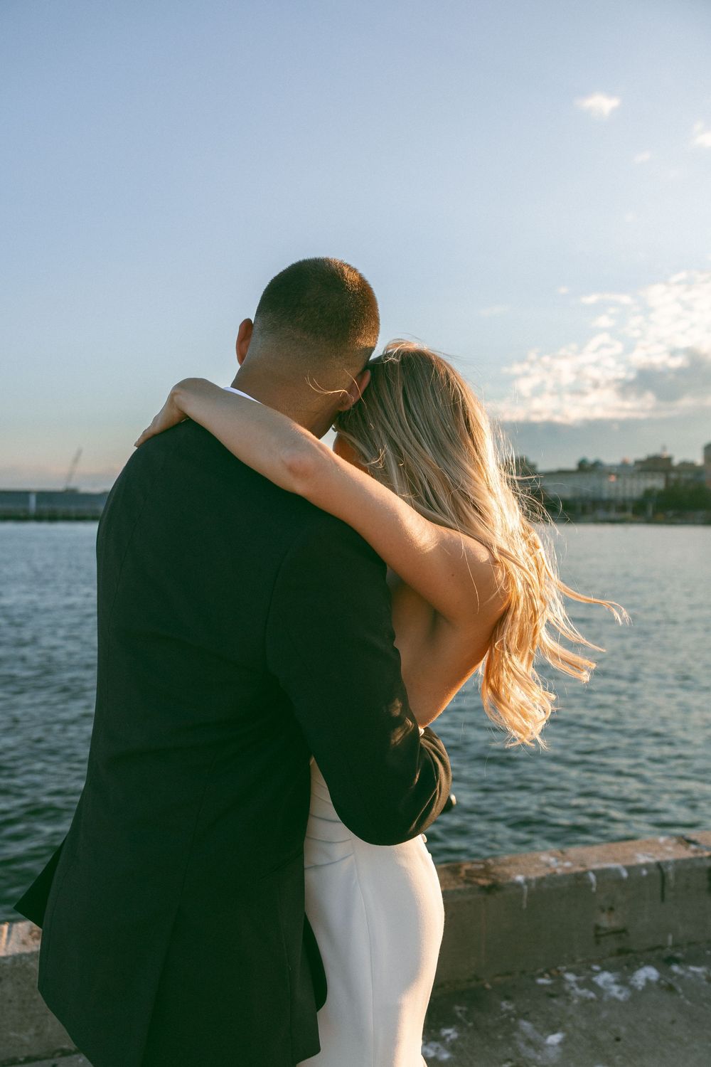 Couple embracing by the waterfront at sunset, woman in white dress and man in dark suit.