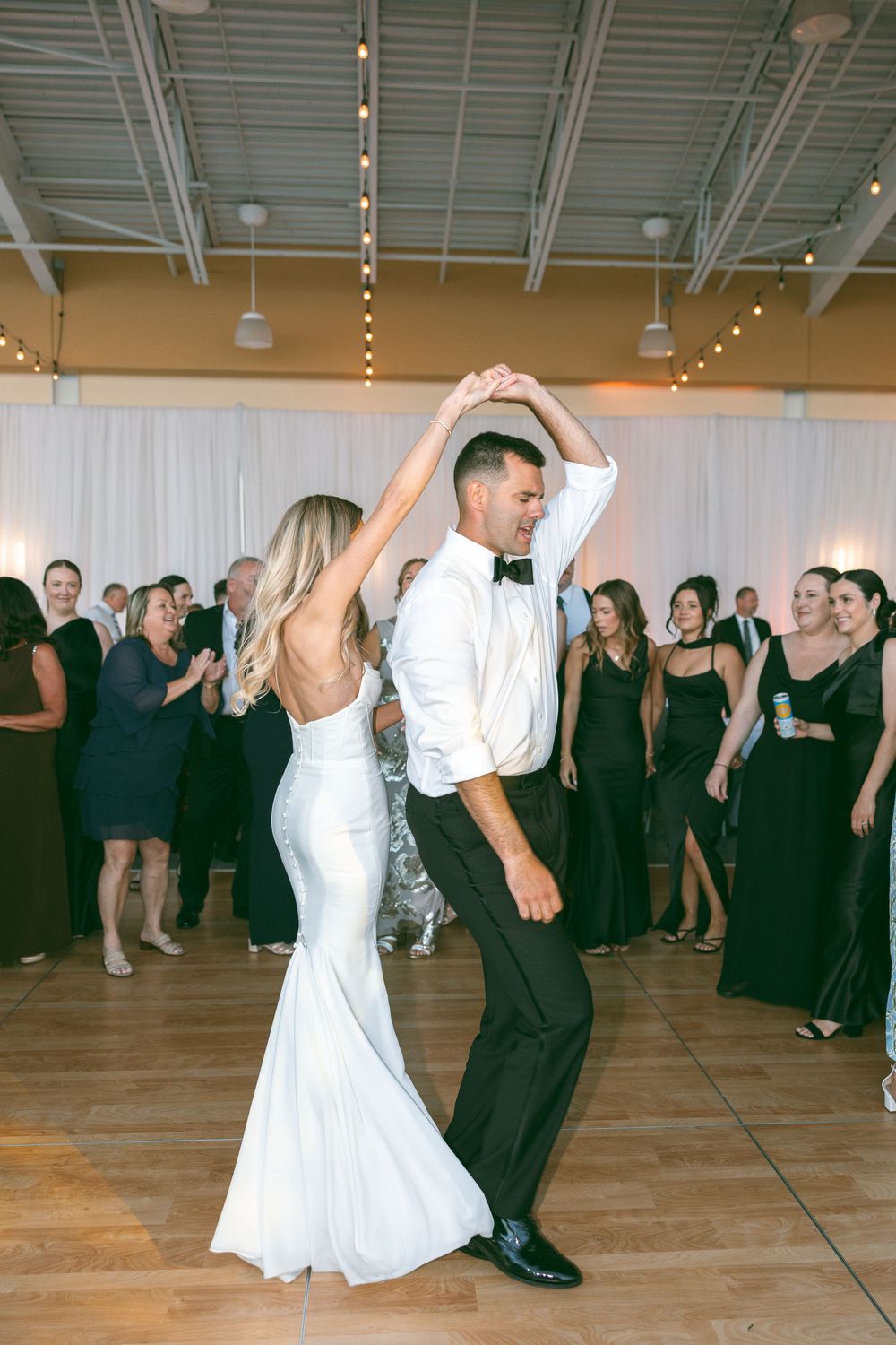 Bride and groom share first dance at wedding reception, guests in black attire watching in elegant venue.