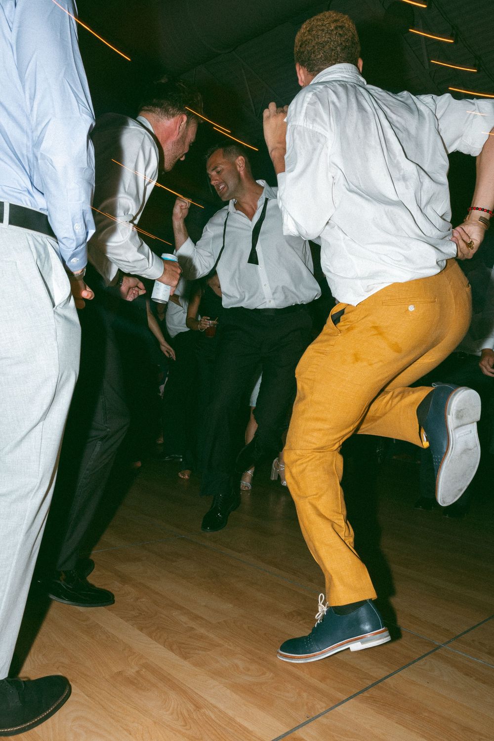 Energetic wedding guests dance on wooden floor, one wearing bright yellow pants and navy blue boots at reception party.