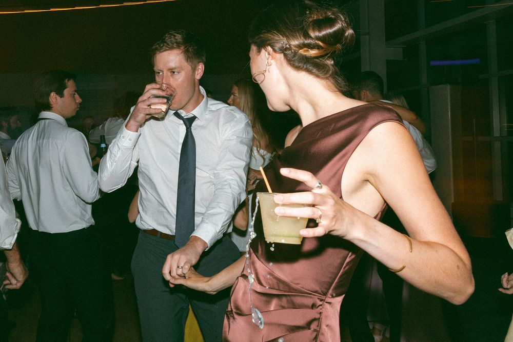 Couple dancing at wedding reception, man drinking from glass, woman in satin dress holding cocktail.