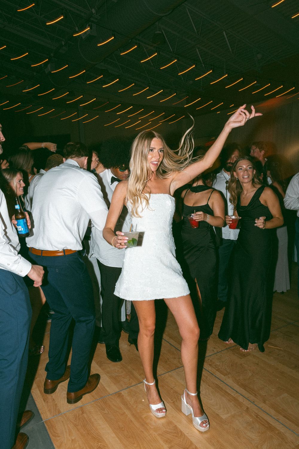 A bride in a white mini dress dances joyfully at her wedding reception, holding a cocktail among celebrating guests.