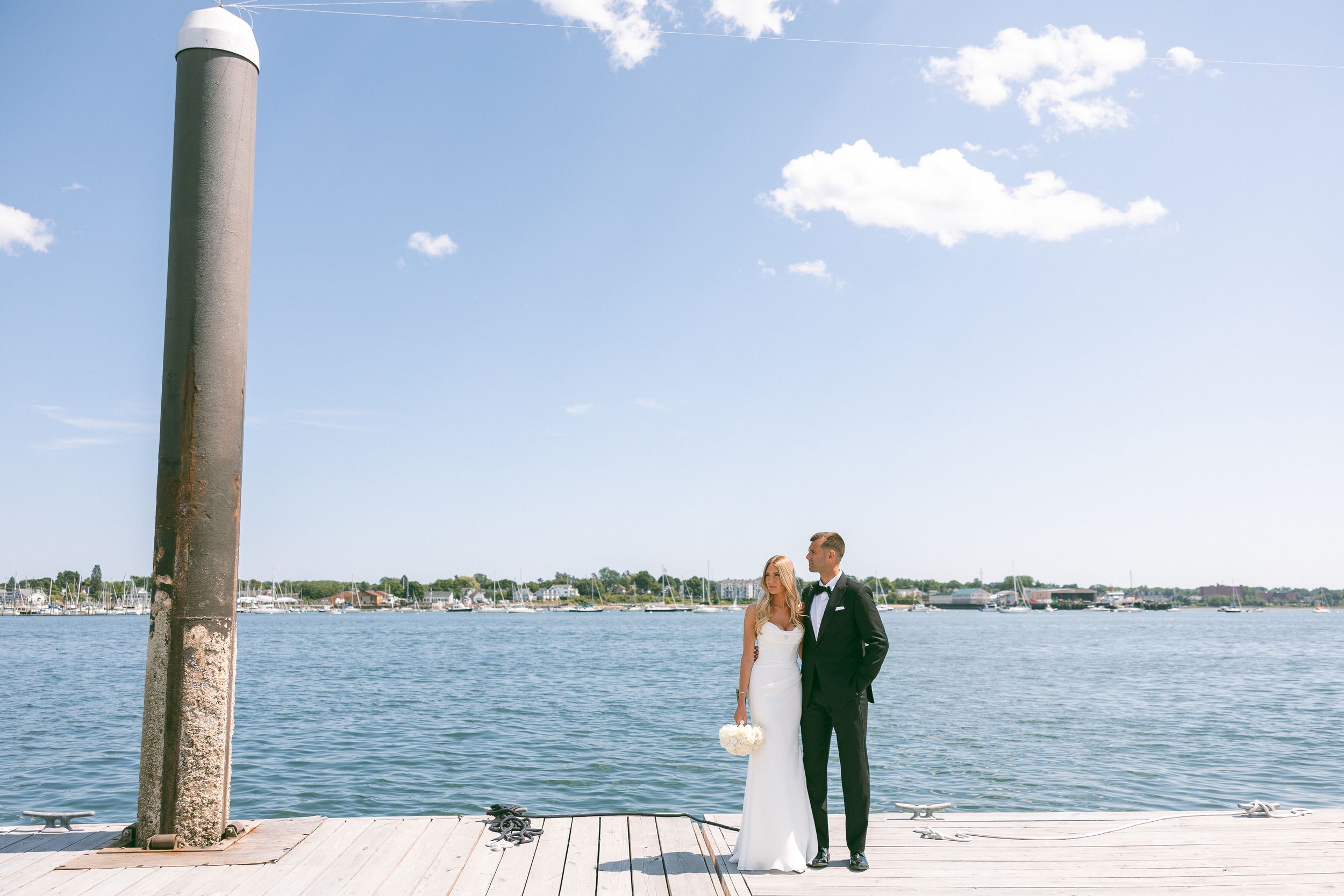Bride and groom share a romantic kiss on a wooden dock by the water on their sunny wedding day.