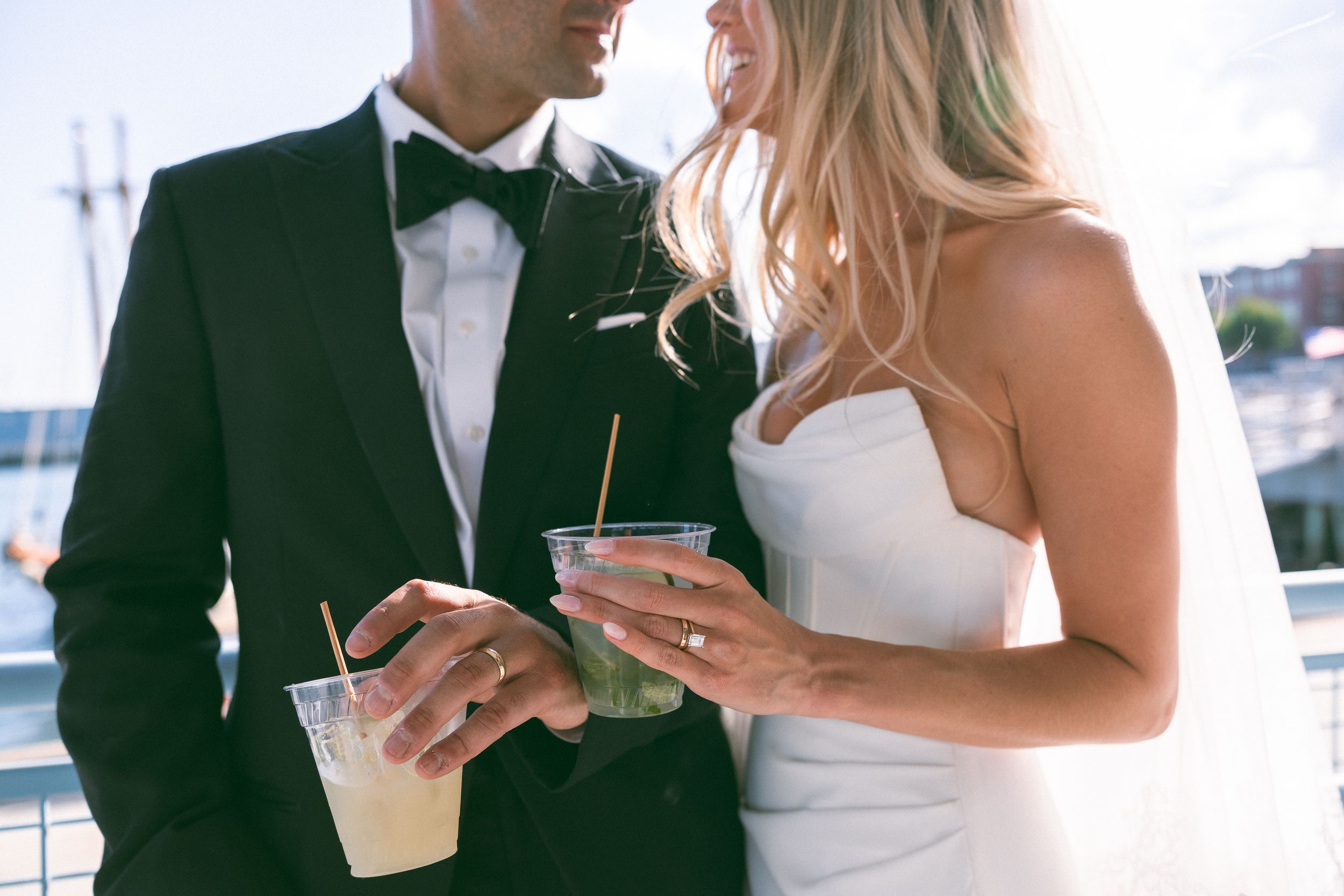 Bride and groom holding cocktails in plastic cups at outdoor wedding reception near waterfront.