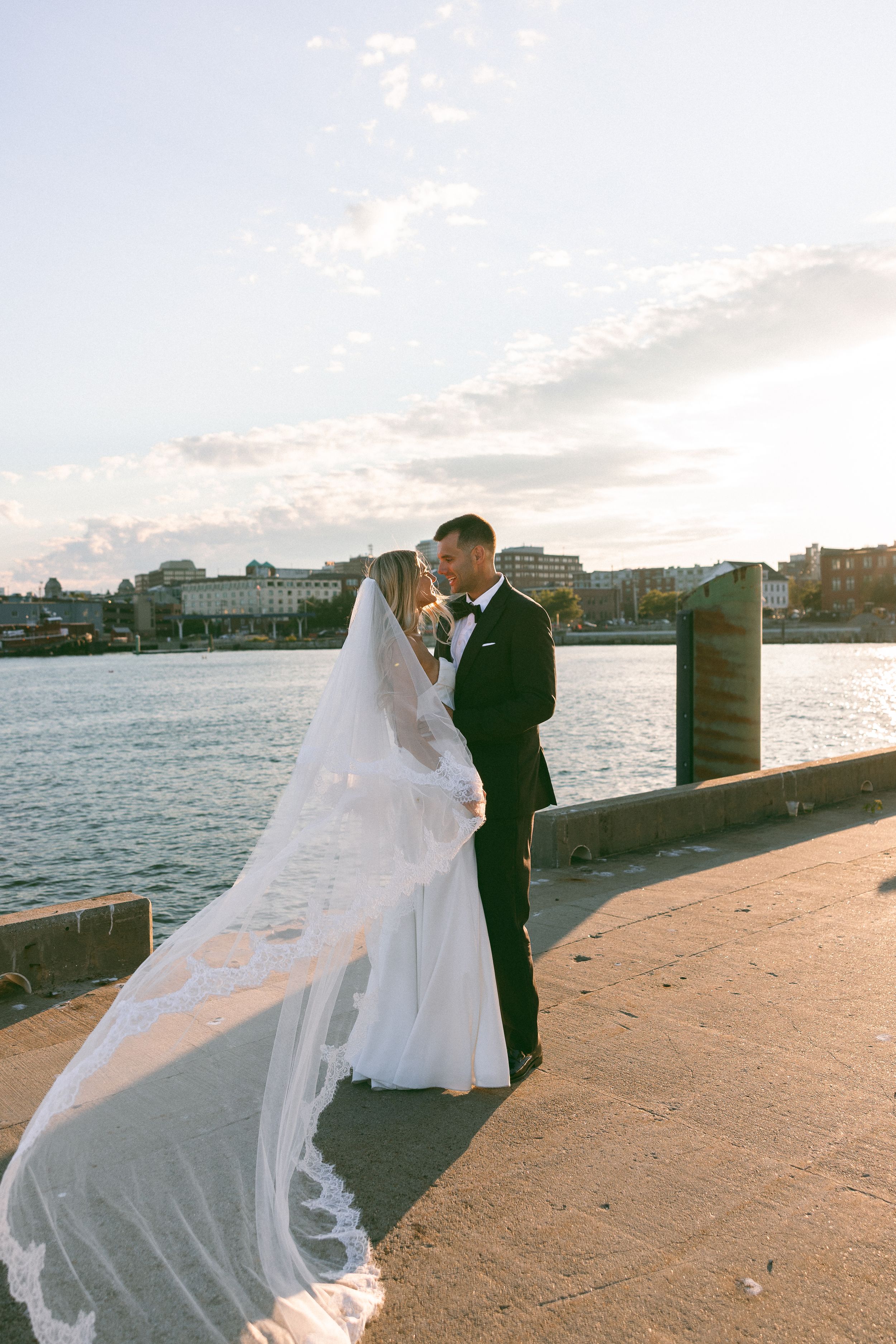 Bride and groom share romantic moment on waterfront pier at sunset, veil flowing in the breeze.