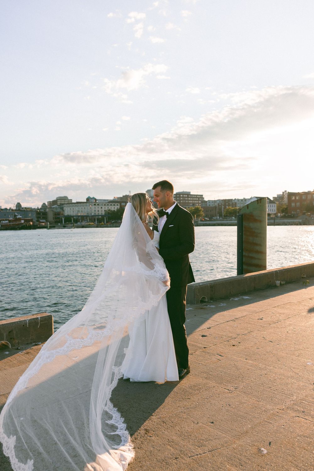 Bride and groom share romantic moment on waterfront pier at sunset, veil flowing in the breeze.