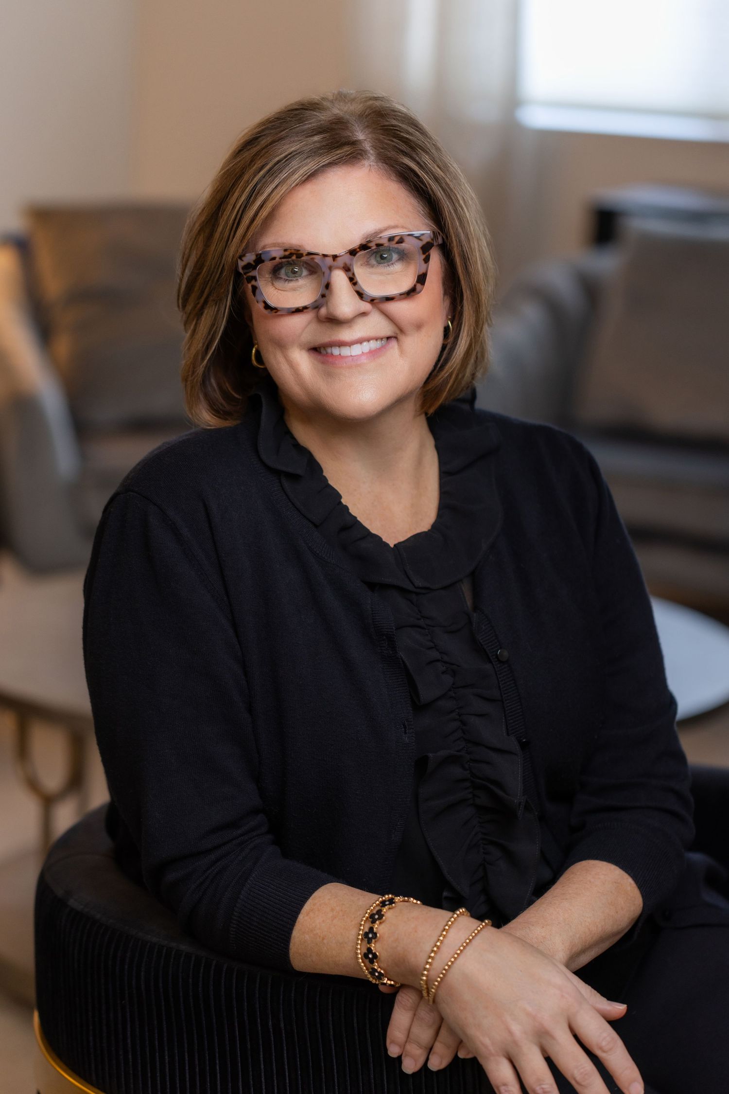 Professional portrait of someone wearing glasses and a black ruffled top sitting in an office setting with a warm smile.