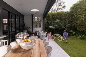 Modern outdoor patio features a sleek wooden dining table under a covered pergola with skylight opening to a green lawn.