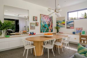 Modern dining area with round wooden table, white chairs, and colorful artwork displayed on white walls.