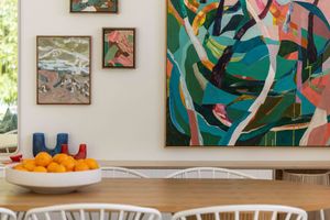 Colorful abstract art pieces hang on a white wall above a table with fresh oranges in a modern gallery setting.