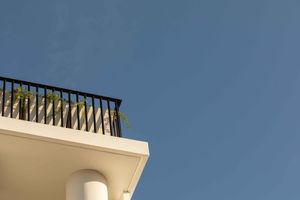 Looking up at a white balcony with black metal railing against a bright blue sky.