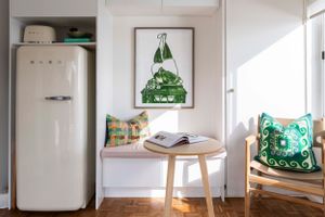 Retro white refrigerator next to small table with modern green mountain artwork and decorative cushions.