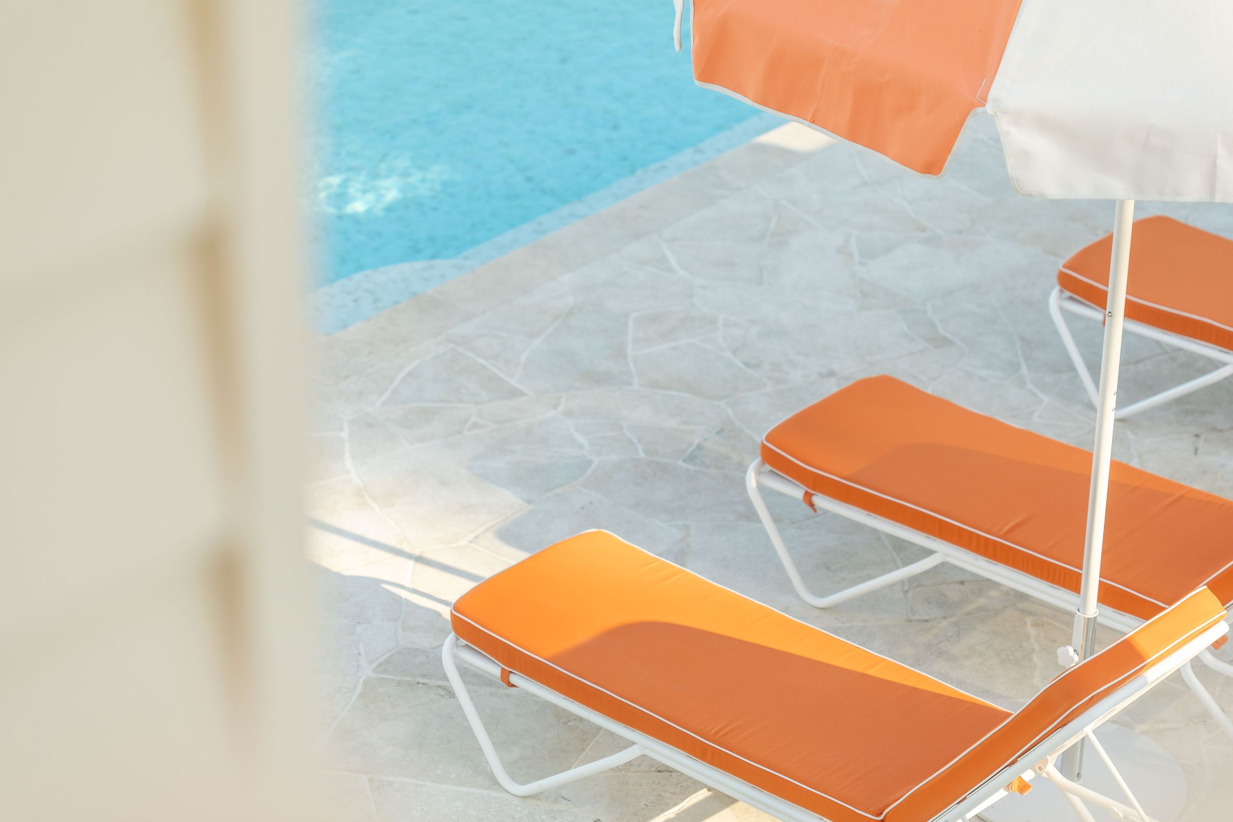 Orange lounge chairs and umbrella beside a bright blue swimming pool on a tiled patio.