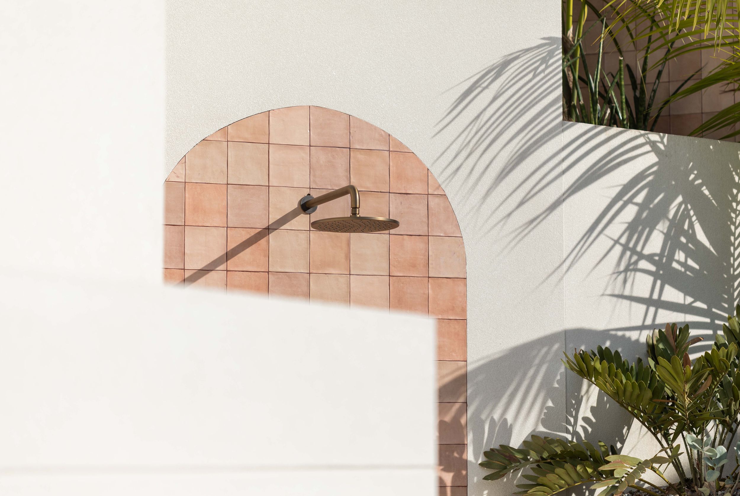 Outdoor shower with terracotta tiles in an arched niche on a white stucco wall, surrounded by tropical palm leaves.