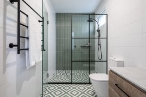Modern bathroom with black-framed glass shower, sage green tile wall, geometric floor pattern, and black towel rack.