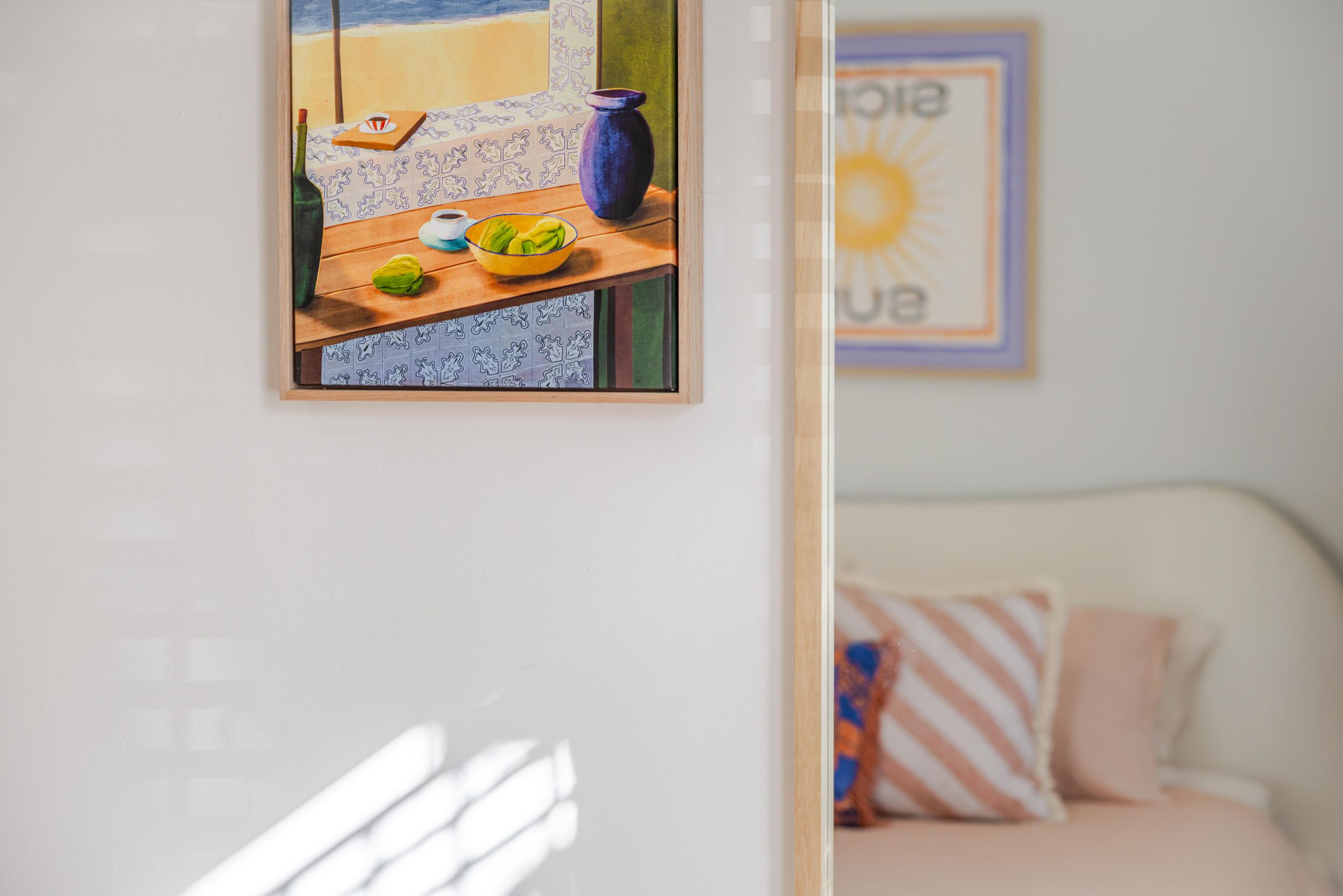 Framed artwork hangs on a white wall near a couch with decorative pillows in a bright, sunlit room.