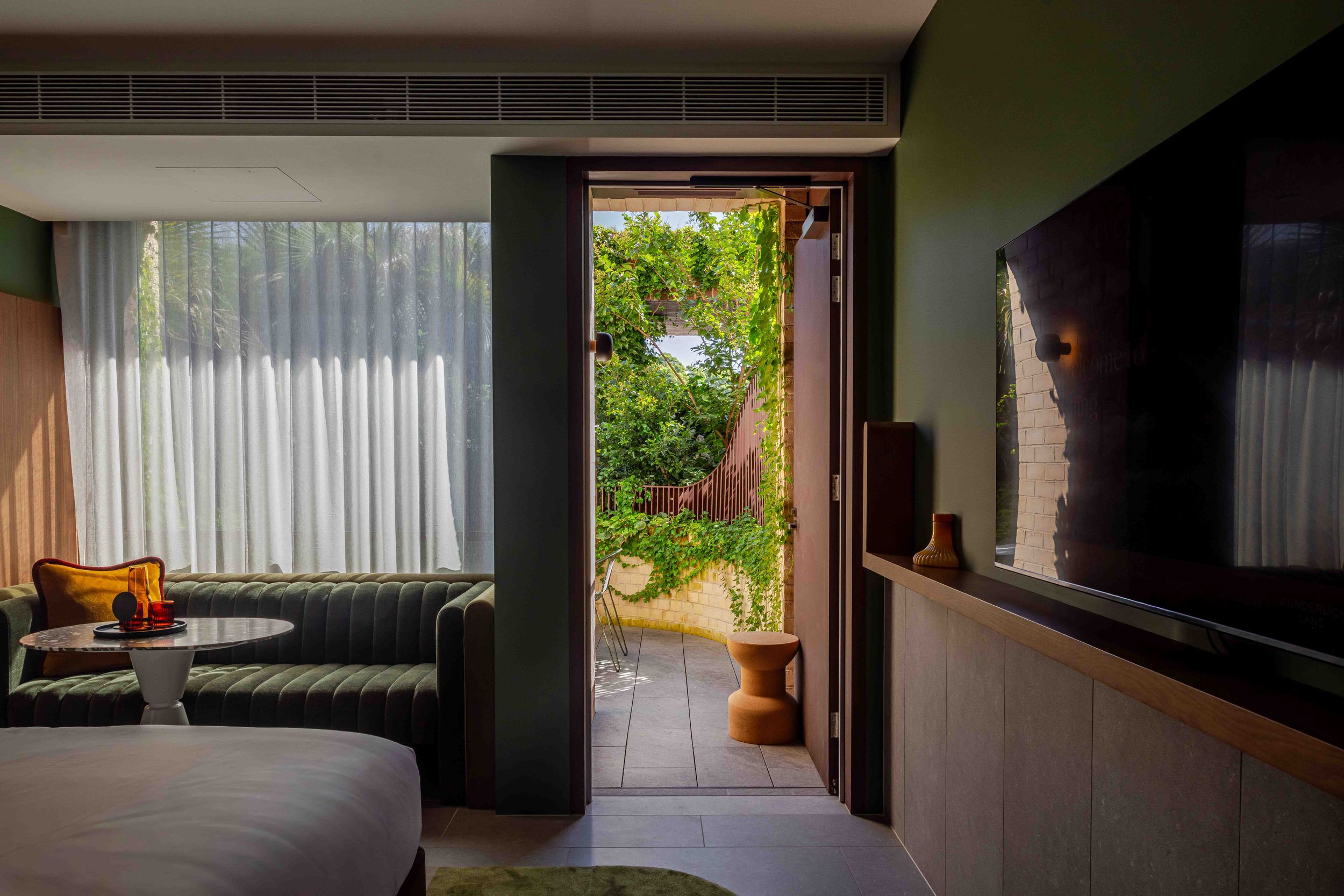 Modern hotel room with green walls, large TV, sofa, and open door revealing lush garden view.