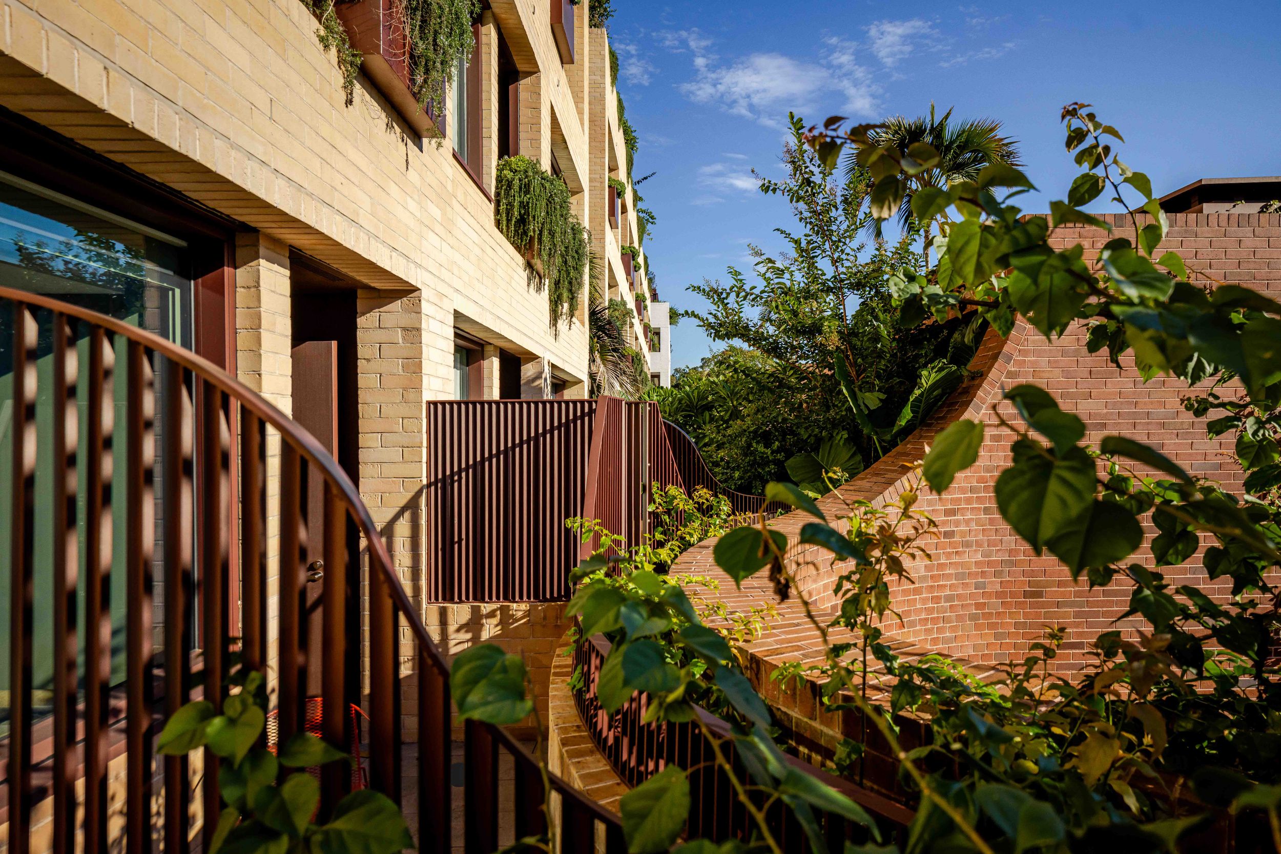 Brick apartment building exterior with lush green foliage, metal fencing, and warm sunlight on a clear day.