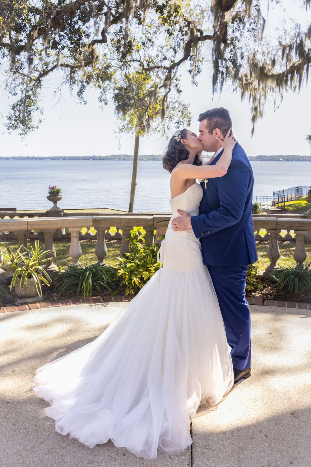 Newlyweds share a romantic kiss on a waterfront terrace with Spanish moss draped trees overhead.