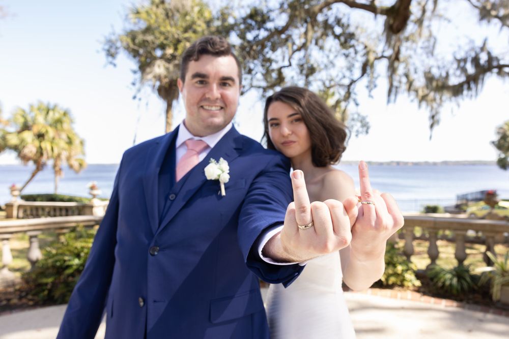 A wedding couple shows off their rings in front of a scenic waterfront backdrop.