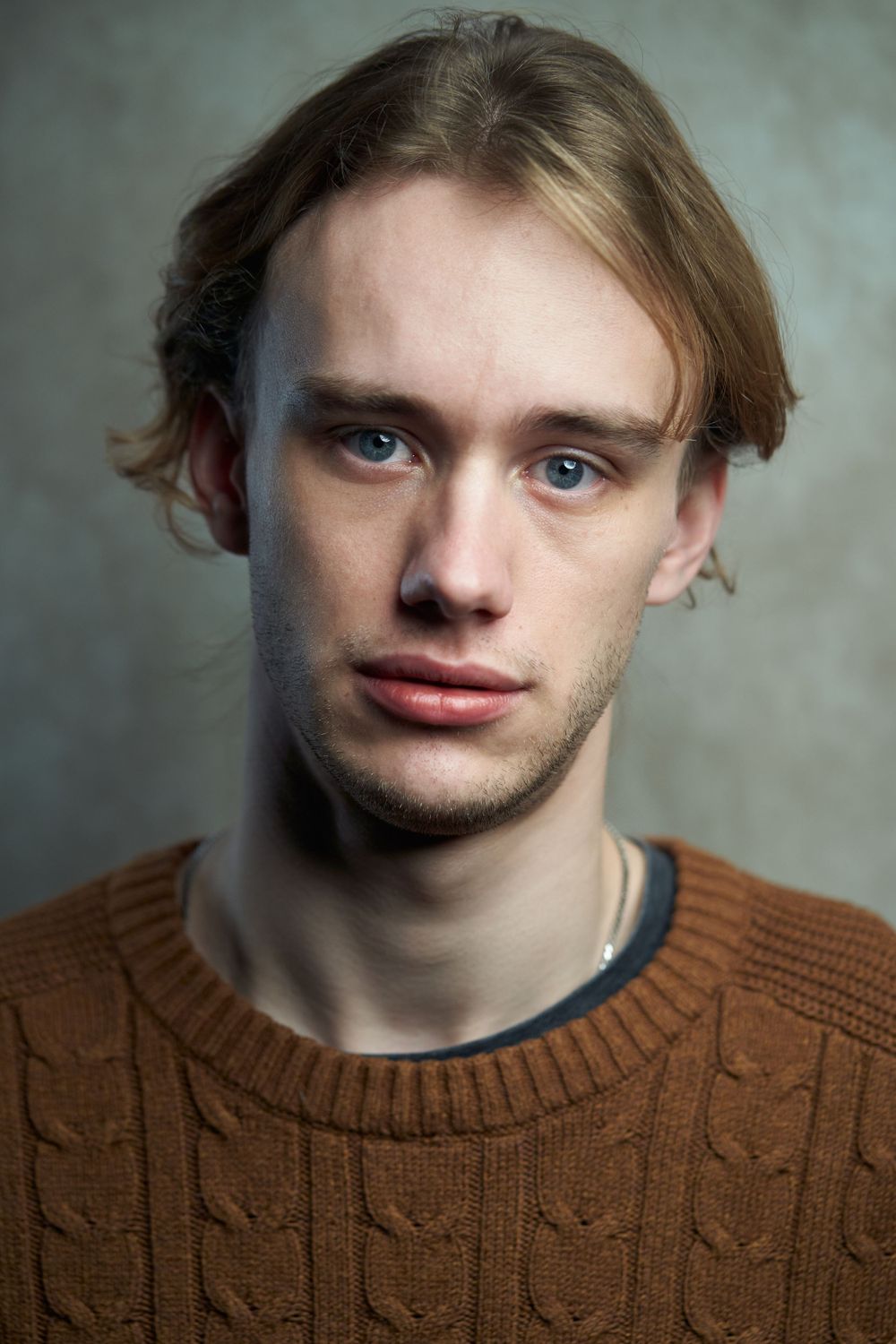 Portrait of a young person wearing a brown knit sweater against a gray background.