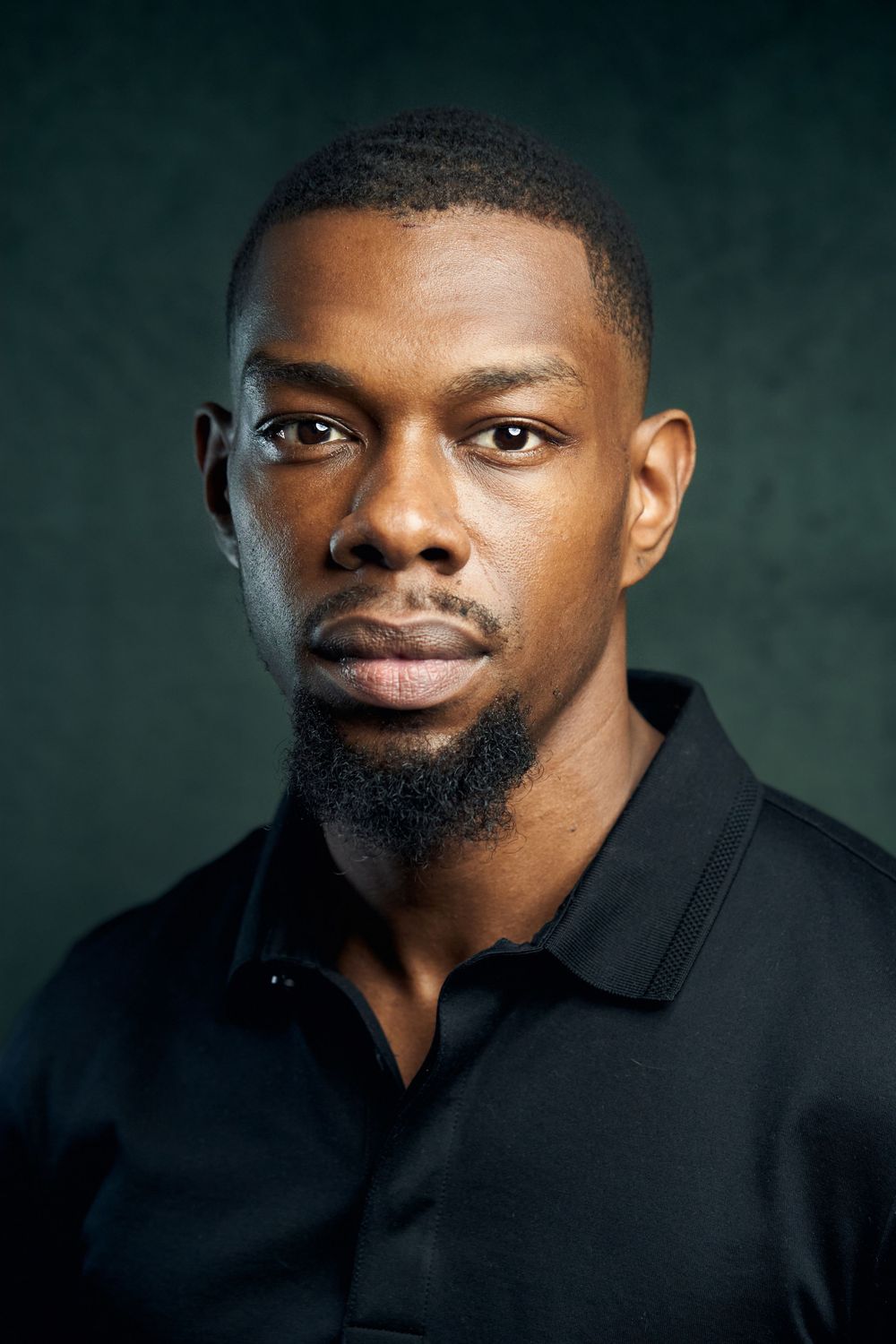 Professional studio headshot of a person wearing a black polo shirt against a dark green background.