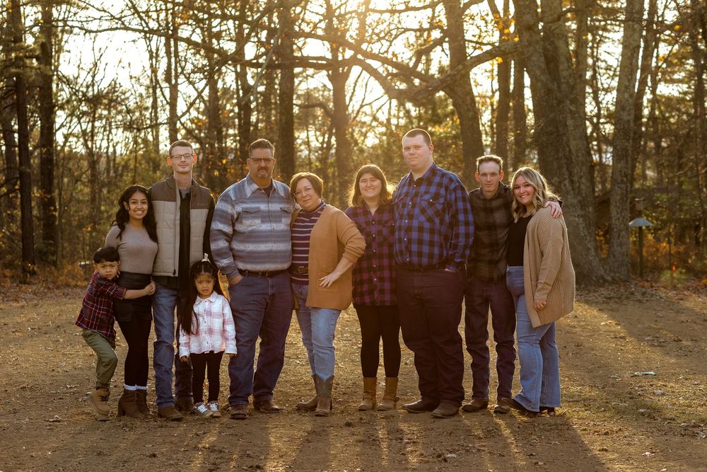 A group of people stand together outdoors at golden hour with sunlight filtering through autumn trees in the background.