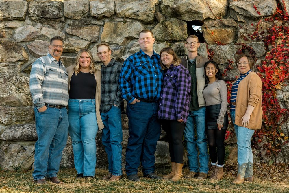 A group stands in front of a stone wall during autumn, dressed casually in plaid shirts and jeans.