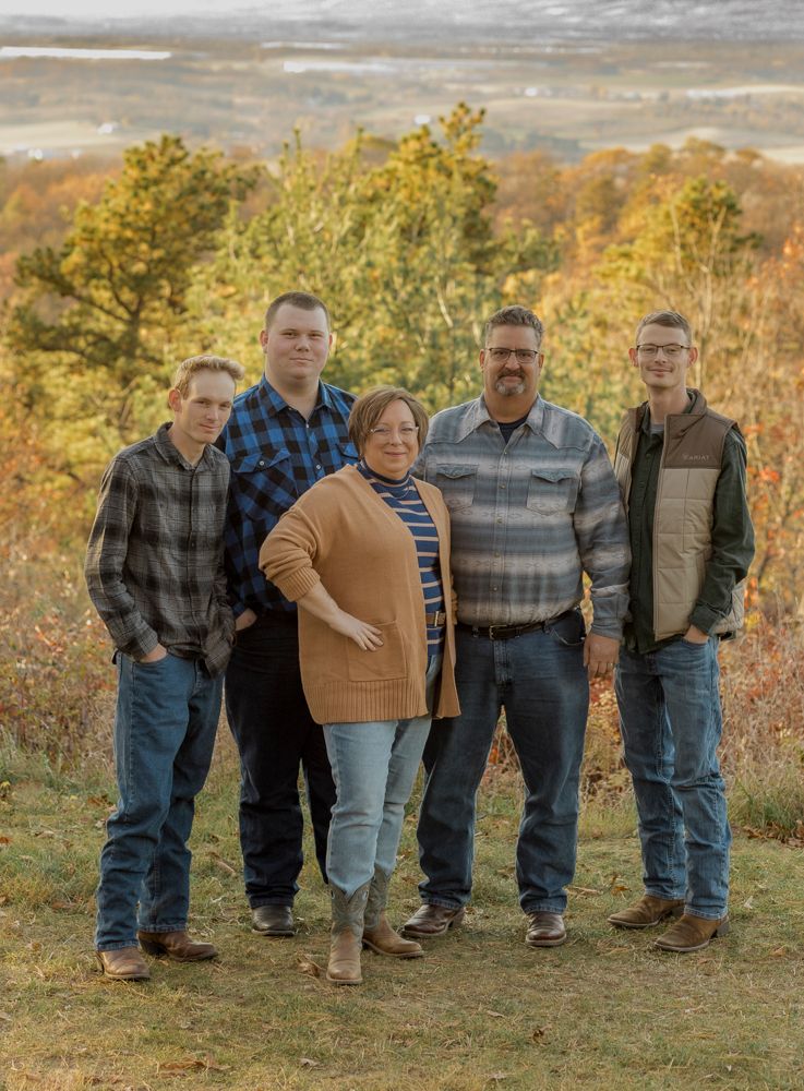 A group poses casually outdoors during autumn with fall foliage and trees visible in the background.