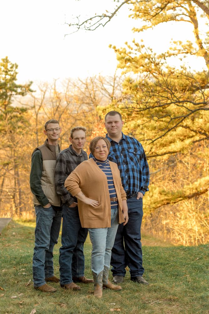 Family group stands together on grassy hillside during golden autumn sunset with trees in background.