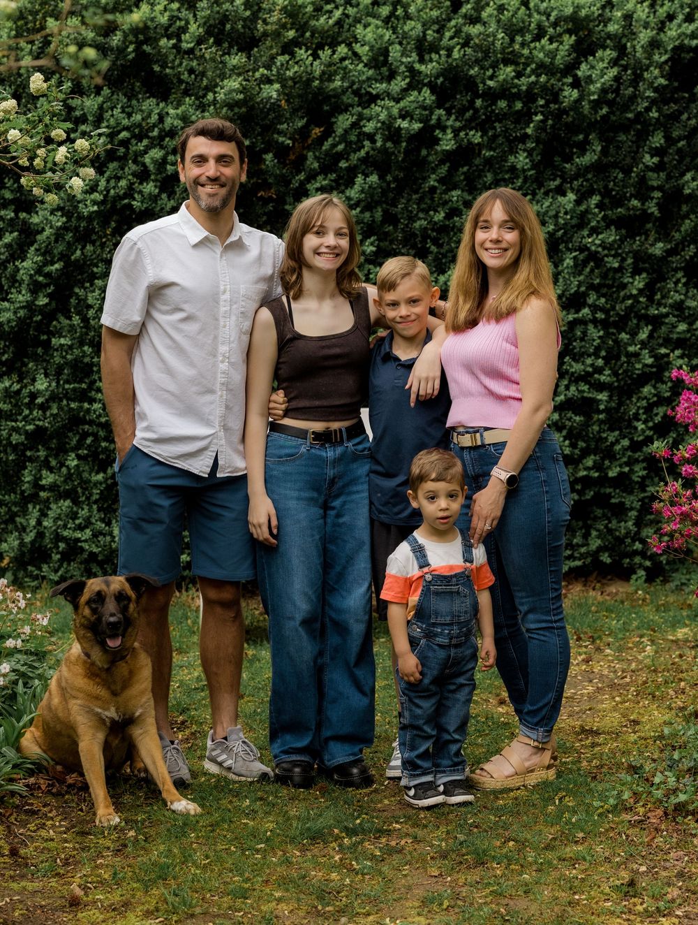 A casual outdoor family gathering with a large brown dog sitting together on grass by green shrubbery.