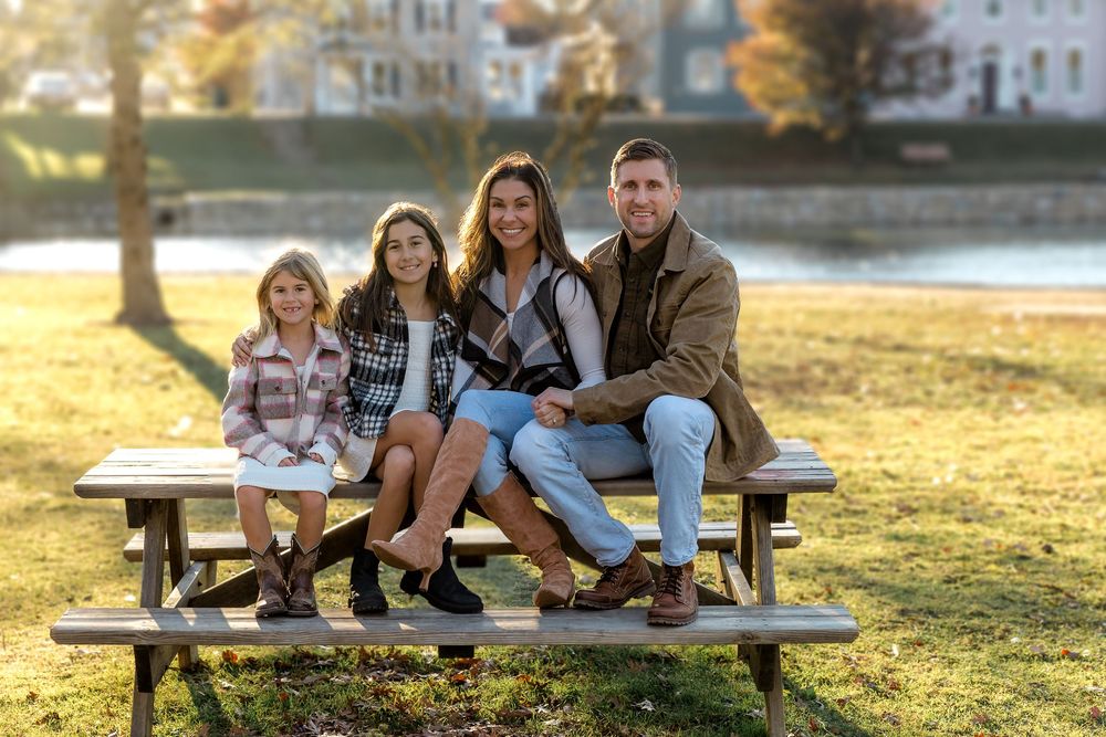 A family sits together on a wooden picnic table near a lake during golden hour on an autumn afternoon.
