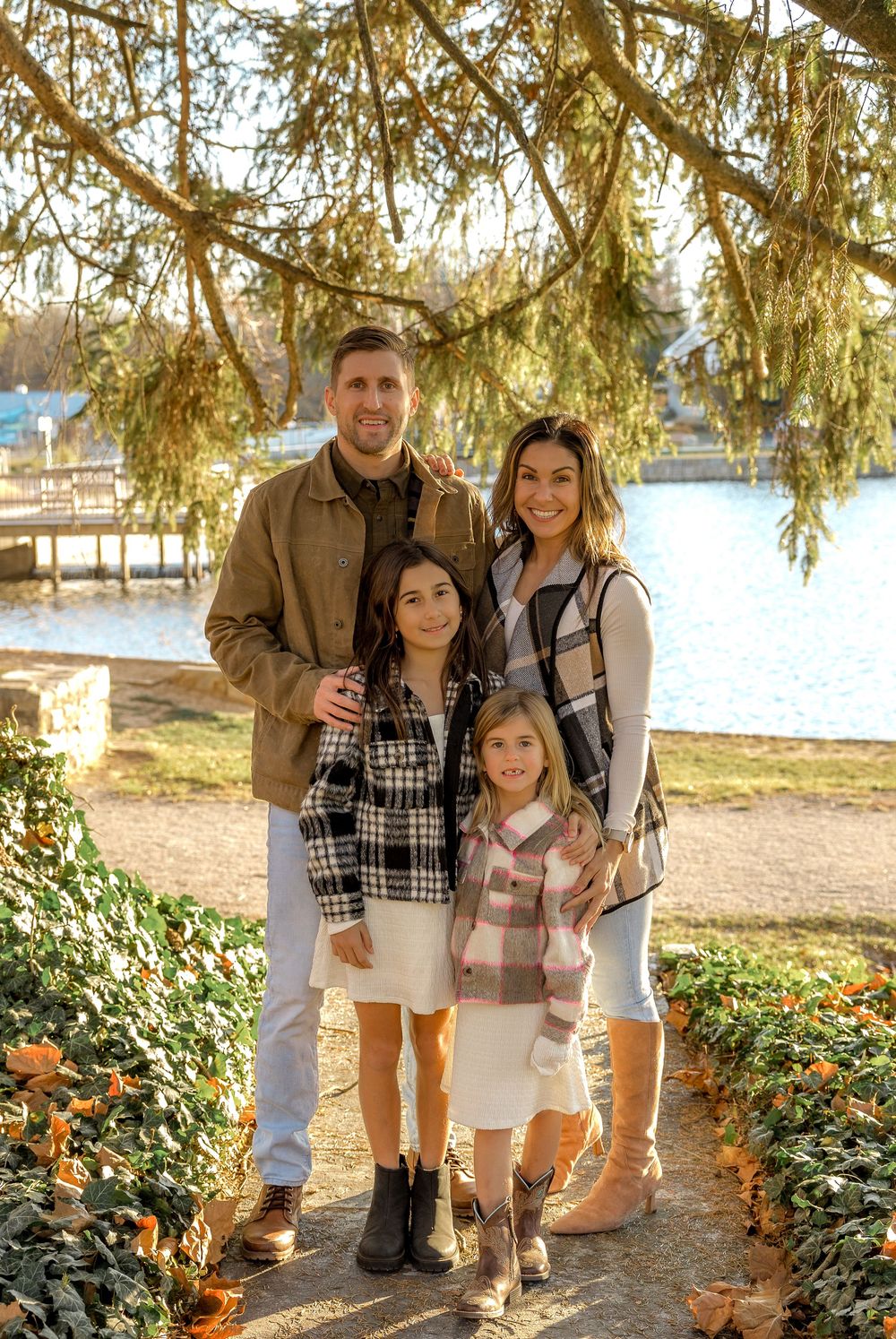 Family in casual attire poses together outdoors near evergreen trees and a lake on an autumn day.